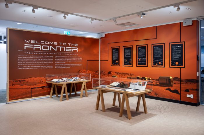 An exhibition space featuring orange walls with Mars landscape graphics, two wooden display tables with glass cases, and text panels including one that reads "Welcome to the Frontier".
