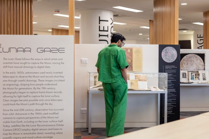 A man in a green matching shirt and pants set stands with his back to the camera, looking at the historical moon documents inside the glass display case.