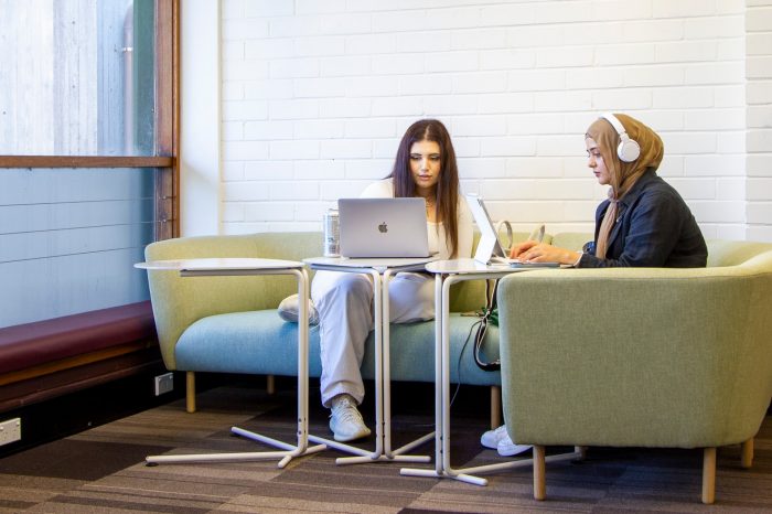 Two students studying on a longer corner couch next to a window. There are laptoptop tables in front of them.