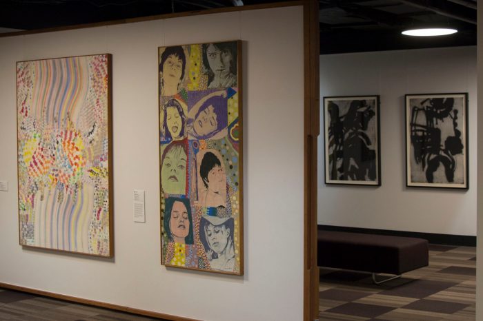 Photo of a gallery space with two brightly coloured framed paintings. The painting in the centre of the frame has a series of woman's faces in repose. In the background is another wall with a two framed black and white paintings. 