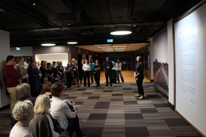 A woman with short dark hair and a pants suit. She is standing in front of the exhibition installation title wall, making a speech. A crowd of guests are looking towards her.