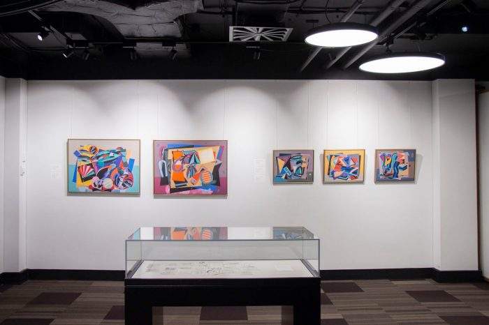 A gallery with five colourful, abstract paintings hanging on the wall. A glass display case is situated in the centre of the gallery space, in front of the paintings. 