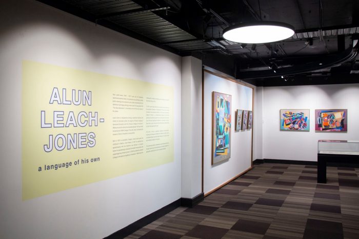 View across a gallery space. On the wall closest to the camera is the title wall, featuring white text outlined in black, with a pastel yellow background. The visible text reads "Alun Leach-Jones: a language of his own". In the distance are various colourful painting mounted on the wall. 