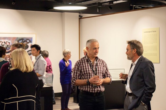 Front view of two guests talking to each other at exhibition opening event, they are holding cups of drinks. Other guests are in view.