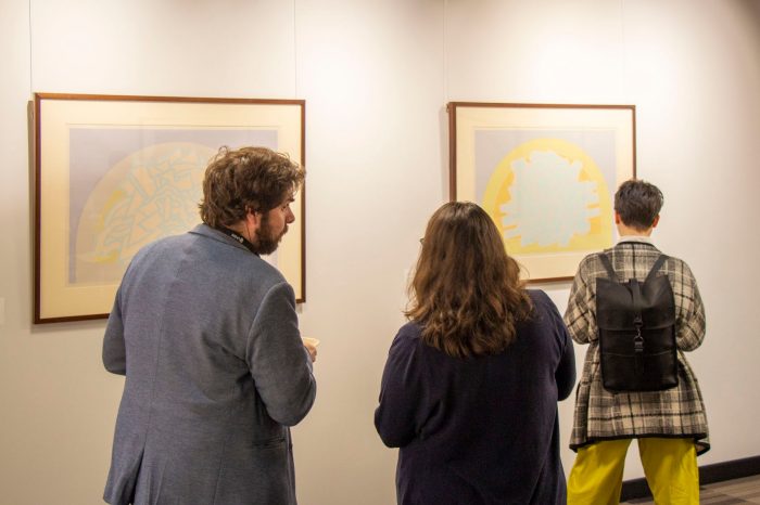 Back view of two guests looking towards an artwork, talking to each other.  Another guest stands closer to one of the artworks.