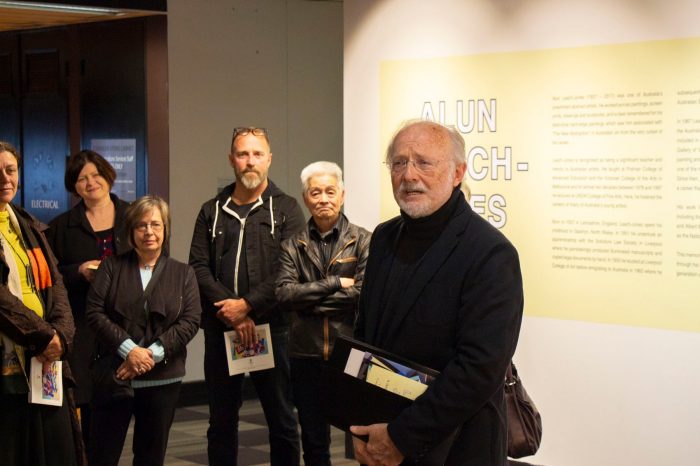 A man with white hair and beard, wearing wireframe glasses and a black suit jacket. She is standing in front of the exhibition installation title wall, making a speech. Guests are looking towards her.