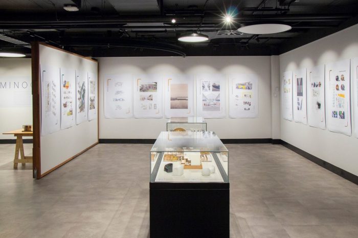 A long-shot photo of an exhibition space, showing three walls, each with four to five posters hanging. To the centre of the photo is a display cabinet, with indiscernible objects behind the glass cover. 