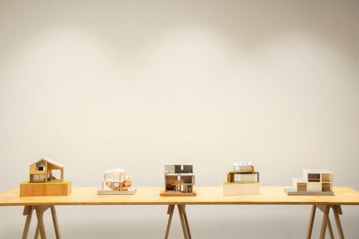 A photo of a wooden table, with five unique three-dimensional architectural models sitting with equal spacing in between. 