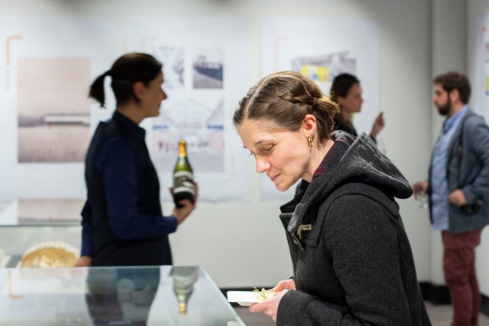 Close-up of a woman gazing into a glass display case. In the background people mingle with drinks in their hands. 