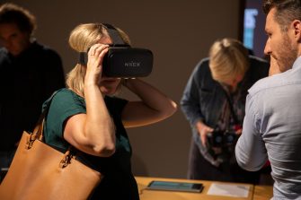 A photo of people attending the exhibition opening event. A lady in a green top wears a VR headset.