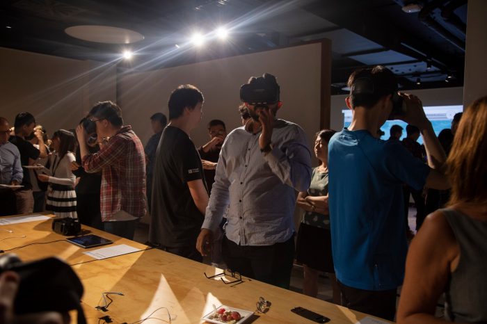 Photo of an exhibition opening event. A crowd of people gather in a gallery space, some of the are wearing VR headsets, others are socialising.