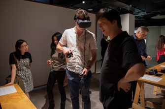Photo of several people gathering in a gallery space. One man is wearing a VR headset device, another man stands next to him talking. People gather and socialise in the background. 