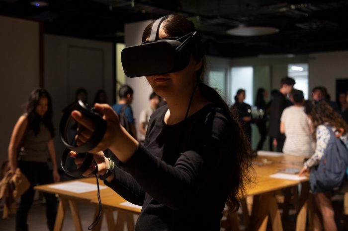 A close-up photo of a lady wearing a VR headset with her arms reached out in front of her. In the background a crowd of people gather and socialise.