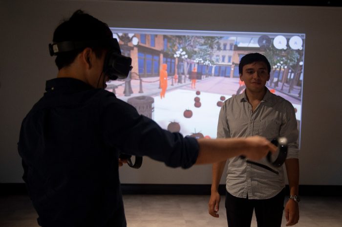 A close-up photo of a man wearing a VR headset, with his arm reaching out in front of him. Another man watches and assists. In the background is a video projection of a 3D virtual reality scene.