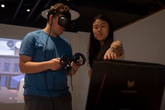 Close-up photo of a man in a blue shirt wearing a VR headset. Next to him, a woman points to a screen that is directly in front of them. 