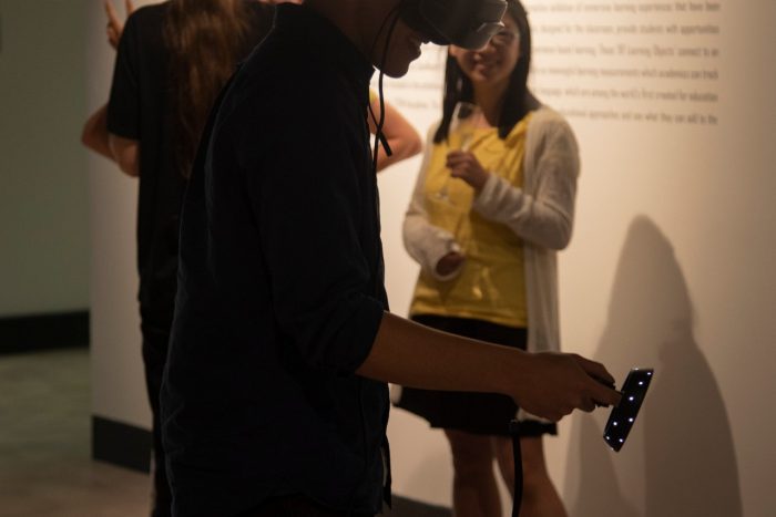 Photo of a man wearing a VR headset, with one arm in front of him. In the background people gather watching him and socialising. 