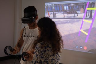 Photo of a man wearing a white shirt in a VR headset. Next to him, a woman with long curly hair watches his movements. Behind them is a video projection of a 3D scene showing an outdoor plaza with a luminescent yellow and purple ladder.