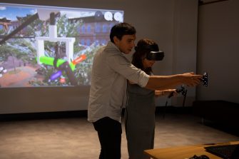 Photo of a woman wearing a VR headset, with her arm stretched out in front of her. A man wearing a grey shirt stands close by, with his arm stretched out near hers. Behind them is a video projection showing 3D scenery.