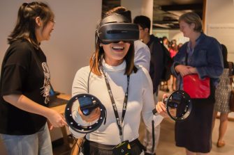 A smiling woman in a white top wears a VR headset and holds two hand-held VR devices in her outstretched hands. Behind her are several people socialising. 