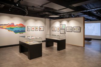 Two glass display cases sit in the centre of a room, a rainbow-coloured line chart on left, artworks in black frames hang on the right.