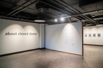 A gallery space with the words "About Closed Doors" across a white wall. Framed artworks can be seen in the background.