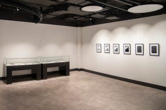 A gallery room with two display cabinets against a wall. Mounted on the adjacent wall are six framed artworks.
