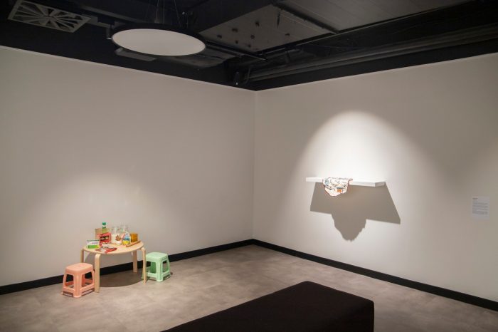 A wide-angle photo of an exhibition space. On the floor to the left are a children's table and chair set. On the wall in a white shelf with cloth hanging over the edge. 