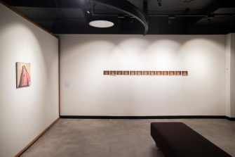 A wide-angle photo of an exhibition space. To the left is a wall-mounted painting, front-on to the camera is a series of red and yellow images lined up across the wall. 