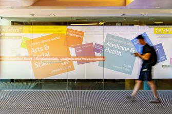 A glass wall in a foyer area, displaying a projection of colourful square graphics and text. The text displays the names of university faculties, such as "Medicine & Health", "Law", and "Engineering". A man walks past the wall.  