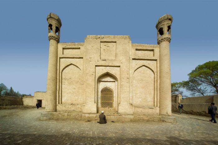 Photo of a sandstone coloured, Persian-style building, with a person sitting at the entrance