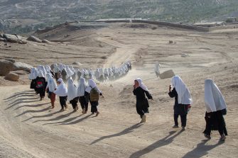 A large group of children dressed in long flowing fabric run away from the camera into a hilly, desert landscape