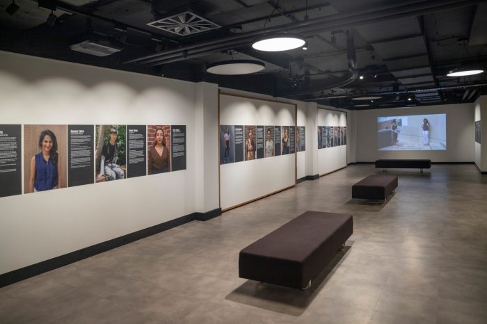 A room with white walls lined with portraits of people interspersed with white text on black panels. A video is projected against the back wall, three benches sit in the centre of the room