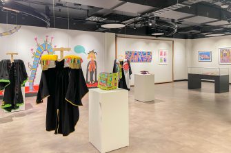 Suspended graduation gowns hang in a room with white walls filled with colourful artworks. Two plinths hold elaborate hats and a glass display case sits near the far right wall.