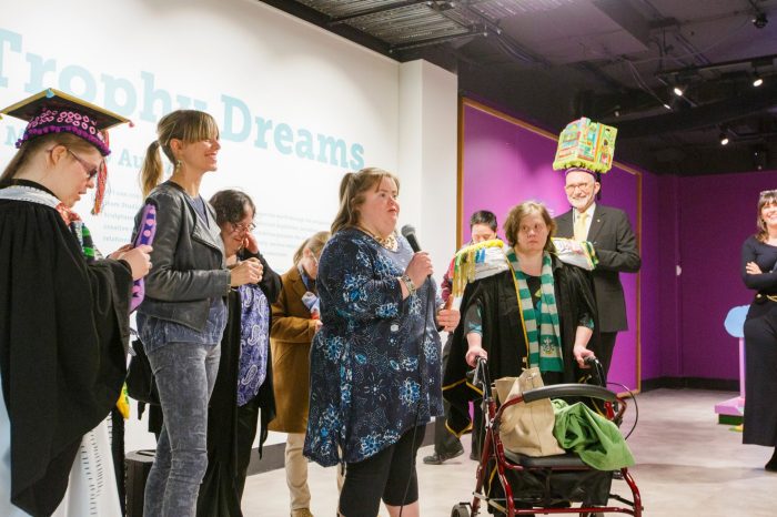 Artist in blue dress speaking to unseen crowd alongside other speakers and artists. Some artists and speakers are wearing or holding artworks.