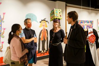 Four guests standing in a semi-circle talking to each other, with a backdrop of exhibition artwork. One guest is wearing a cap from the exhibition.