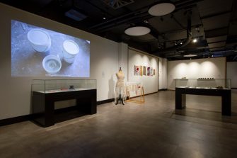 A gallery space with several separate artworks. Artworks include a video projection, objects in two display cabinets, a textile work on a dressmaker’s mannequin, colourful wall-mounted textile works, and sculptural objects on two wall-mounted shelves.