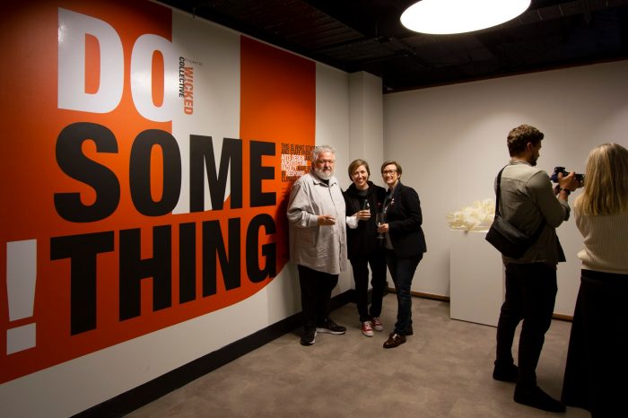 A man and two women standing against the orange title wall of the exhibition.