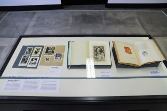 A glass display case contains small multicoloured prints affixed to sheets of paper. Two books supported by grey foam wedges are propped open to pages featuring a colourful, floral bookplate and a series of two black and white bookplates of an urn.