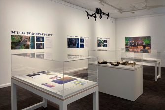 A corner of the Sonus Maris exhibition. The space features white walls and a dark brown carpet. Several display cases are placed throughout the room. There are several prints of aerial photographs showcasing various geographical locations.