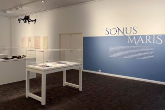 A corner of the Sonus Maris exhibition with a pale-blue wall to the right with the words 'Sonus Maris' in a stylised, uppercase typeface. Several display cases are positioned in the space and a drone hangs suspended from the ceiling.