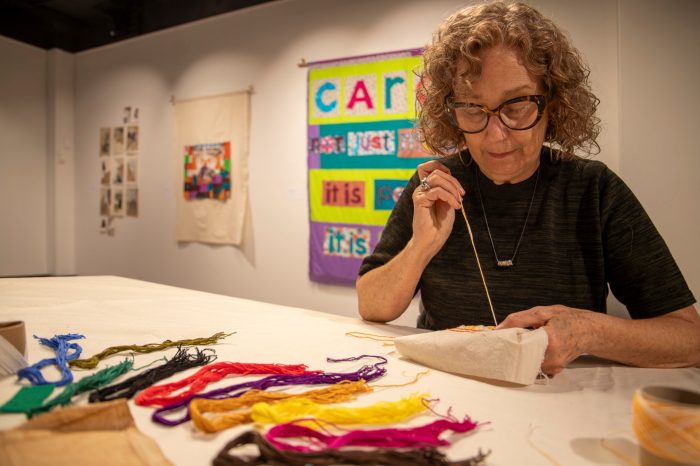 A woman sits at a table sewing with brightly coloured threads gathered in the foreground.