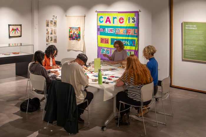 A group of people seated around a white tables sew small squares of fabric with coloured thread.