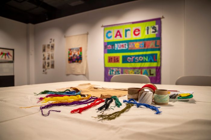 A white table in the foreground contains colourful thread. In the background, the walls are lined with stitched artworks.