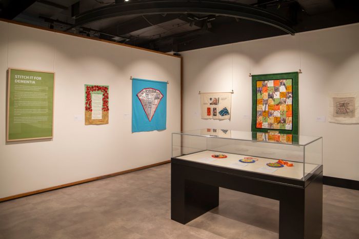 A room with two walls lined with stitched artworks and banners. A display case contains small, colourful round rosettes.