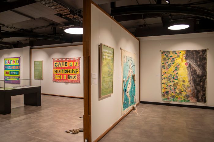 Two rooms containing colourful suspended artworks, some stitched banners others large body maps.