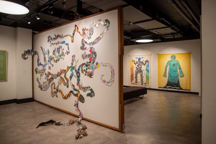 A wall protrudes into the space with a sprawling artwork covering the entire wall featuring twisting tentacles collaged with colourful pictures. In the background against the far wall, two artworks hang featuring simplified human forms.