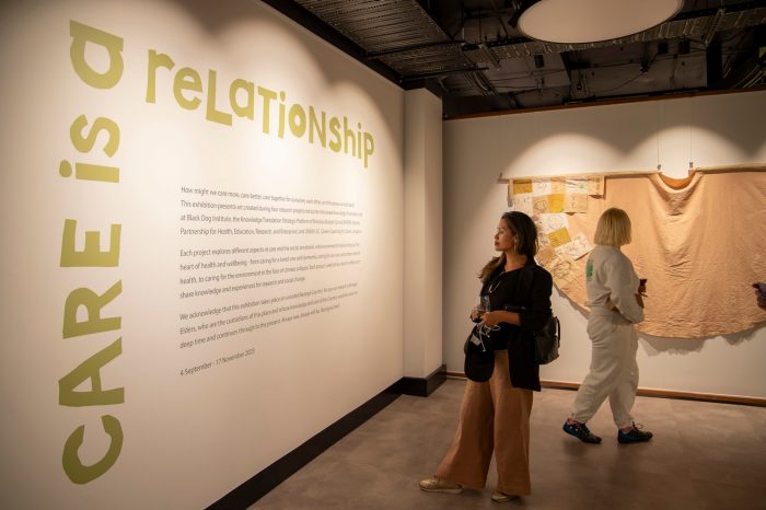 A woman stands reading wall text with the words ‘Care is a relationship’ in green block letters. A woman with her back to the camera looks at a textile artwork suspended on the wall.