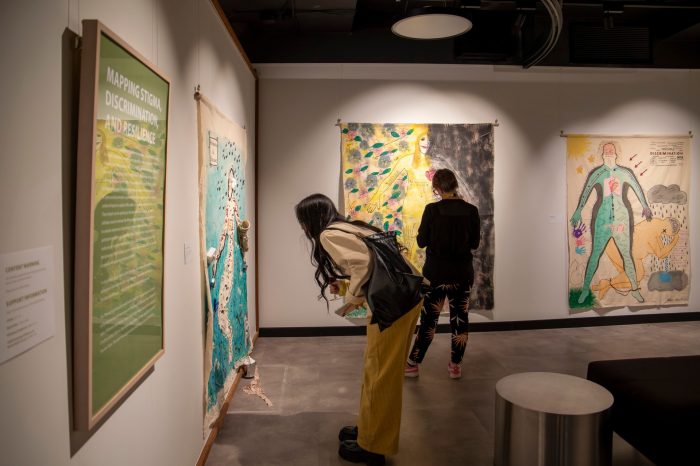 Two women stand in a room reading a green sign with white words and a large body map featuring the outline of a figure against a blue background with found materials covering the surface