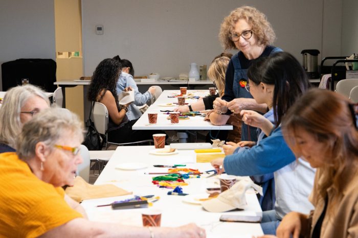 A group of people seated at long white tables sew small squares of fabric with coloured thread.