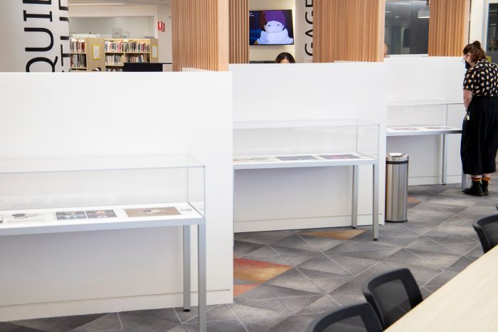 A long shot photo of a library space with three display cabinets against a staggered set of walls. A person is looking at the display cabinet furthest from the camera.